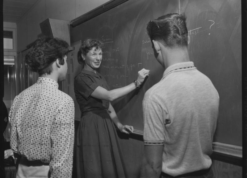 An old back and white photo of a female teacher drawing on chalkboard in front of two students.