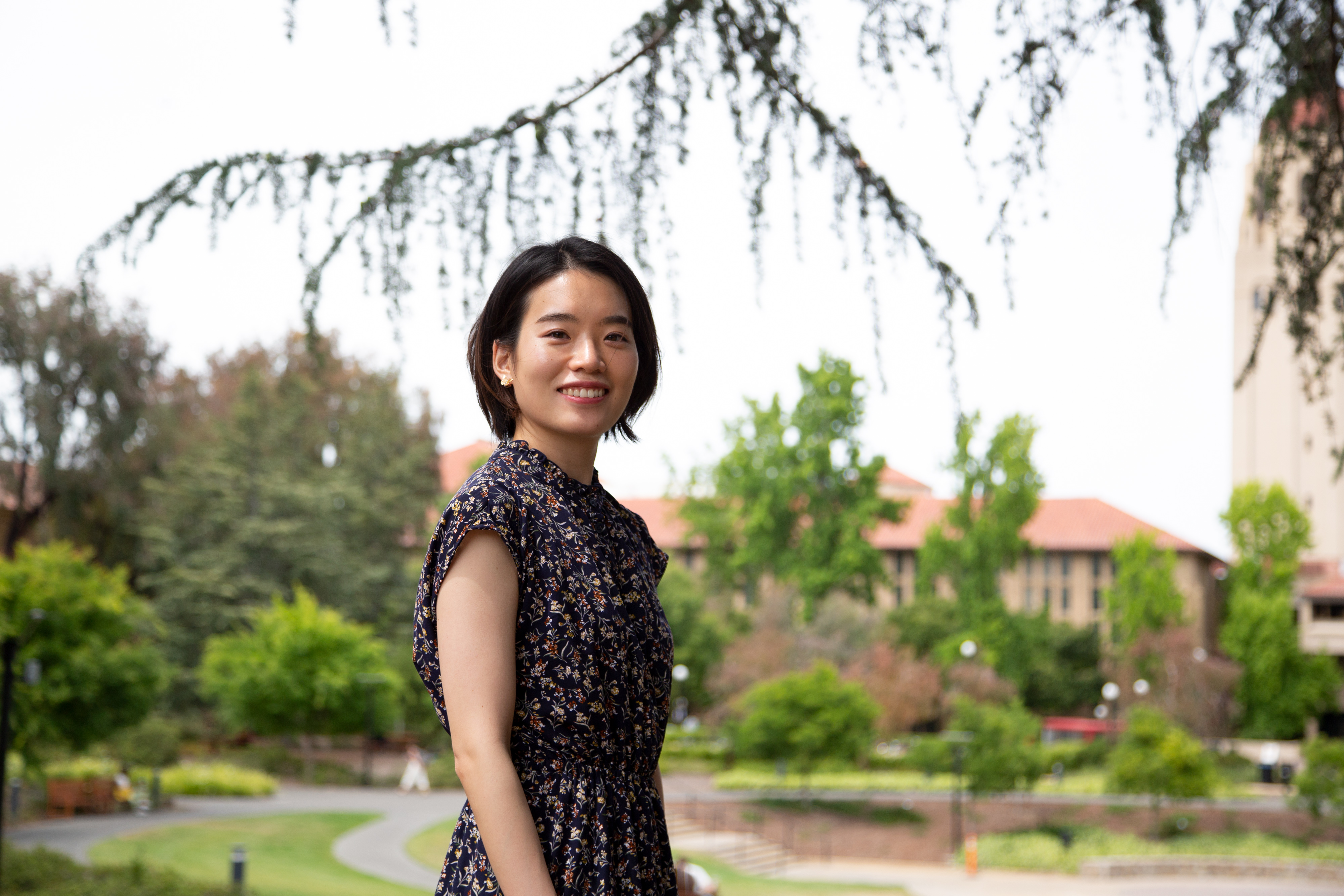 Photo of Risa Ninomiya standing outside on the Stanford campus, with leafy trees and buildings with red tiled roofs in the background