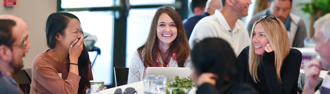 Women smiling at camera at a table full of attendees talking.