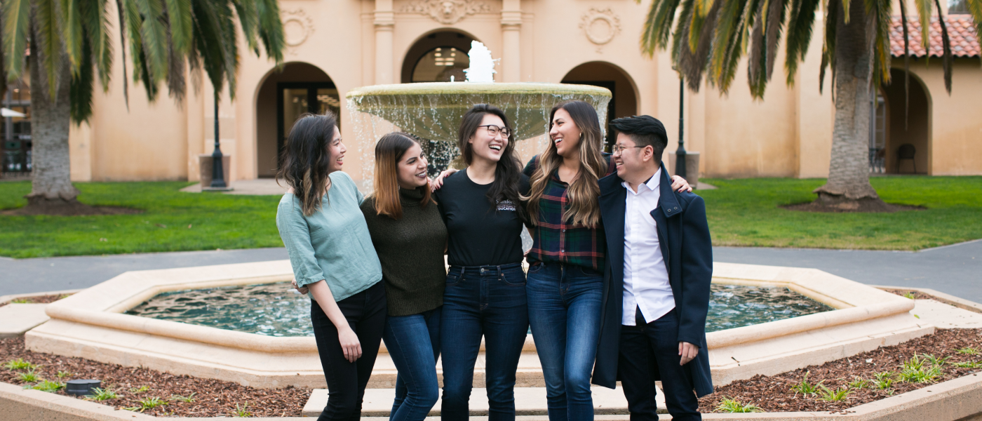 A group of students smiling to each other in front of a fountain.