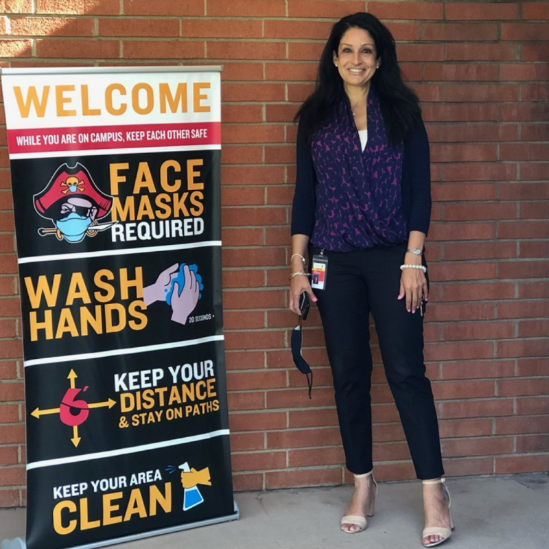 Photo of Facia Desmond at her school, standing near a sign that welcomes students and gives guidance about how to keep others safe during COVID