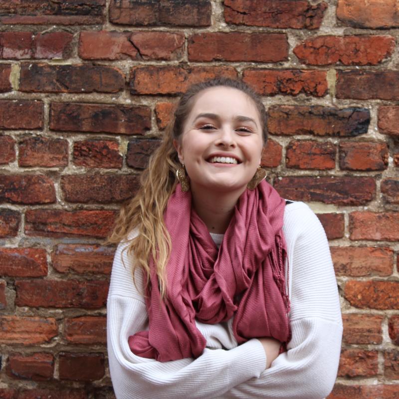 Rachel Herrera, smiling radiantly, arms folded, in front of a brick wall