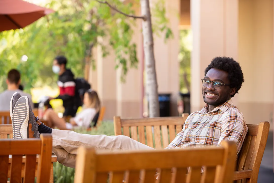 Photo of Cordy McJunkins, sitting on a bench with his legs on a table with some students in background