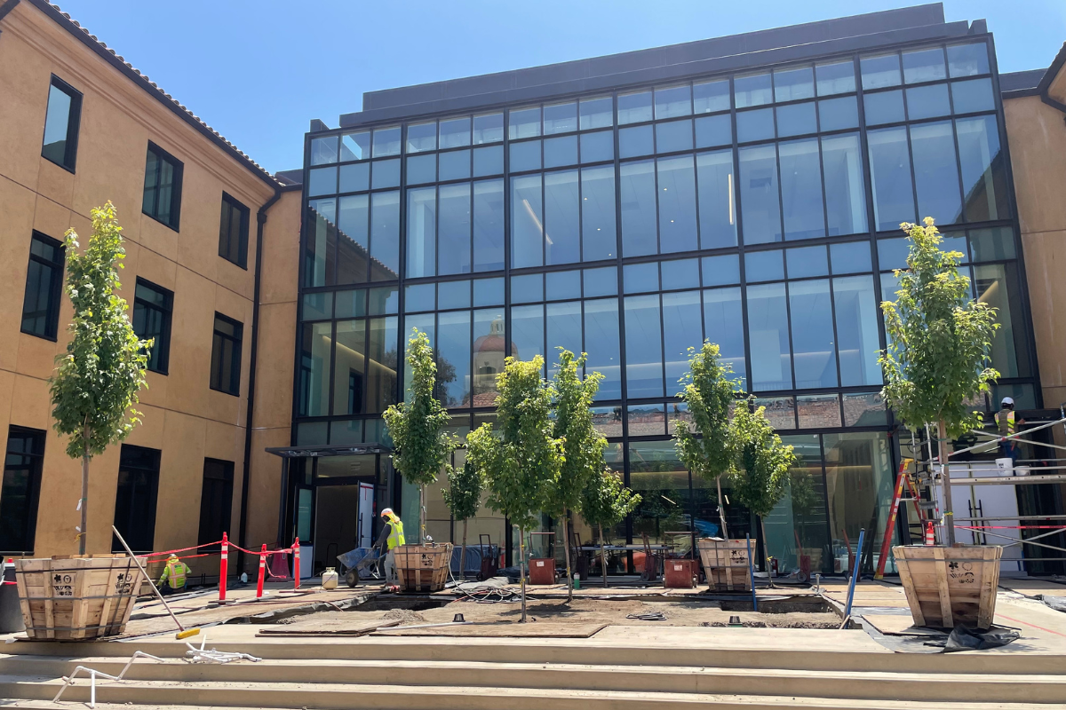 A view of the new building, with its glass wall reflecting Hoover Tower, as trees are being planted in its entry courtyard