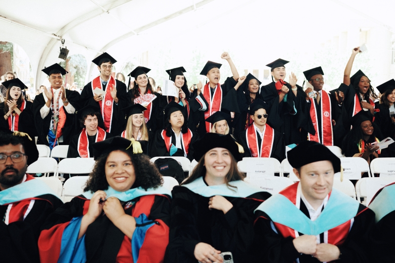 Students in graduation gowns sit in the foreground while another group stands in back. 