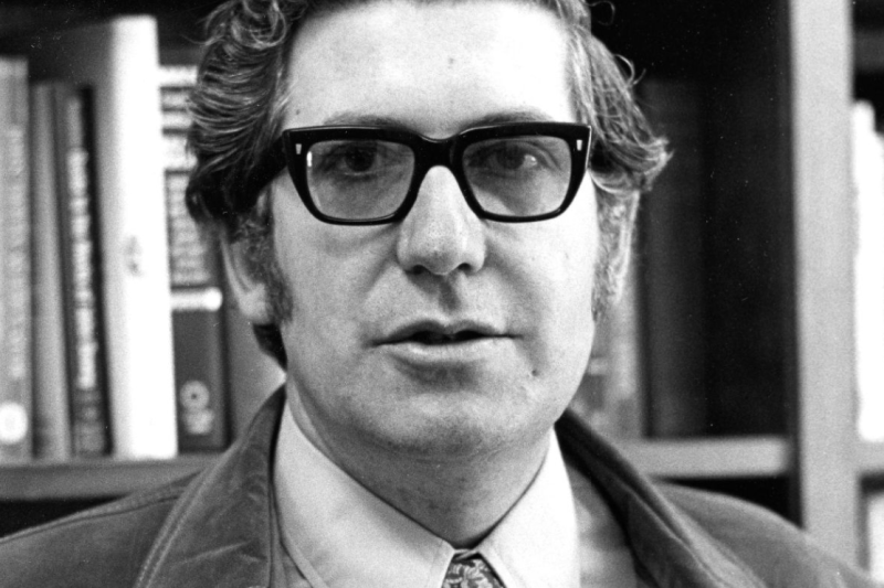 Picture of a man standing in front of a bookshelf wearing a tie and jacket and glasses. (Stanford University Libraries))