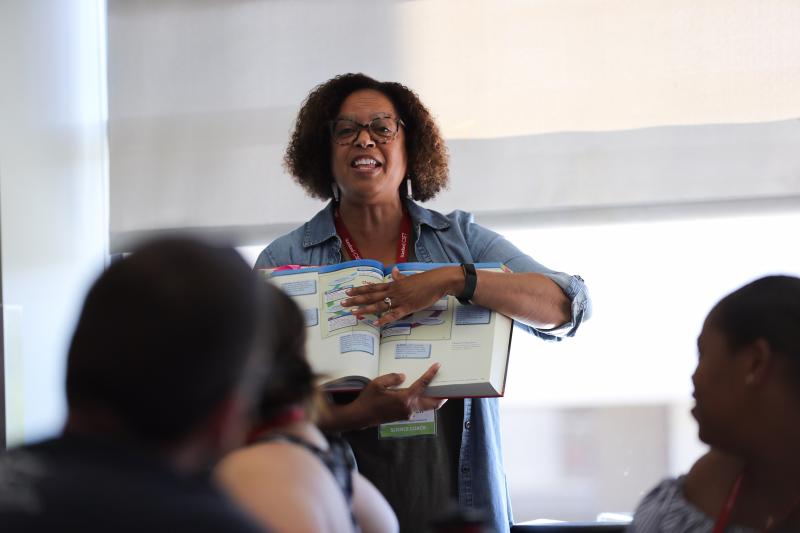 A female teacher in front of the classroom pointing at a line in a textbook.