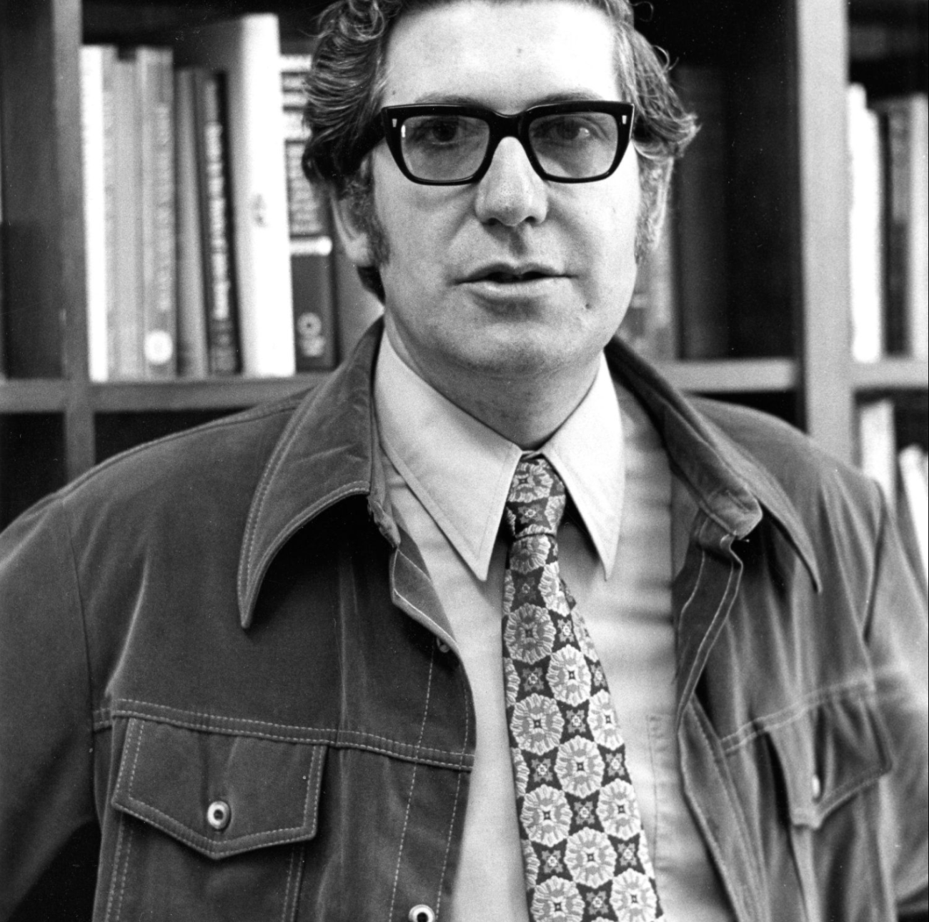 Picture of a man standing in front of a bookshelf wearing a tie and jacket and glasses. (Stanford University Libraries))