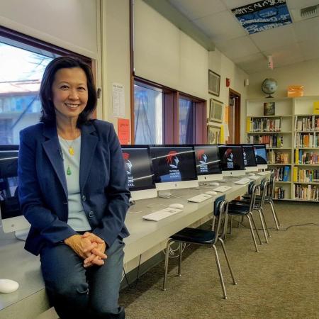 Ting Lan Sun, smiling, in the media center of a school with a row of computers in the background